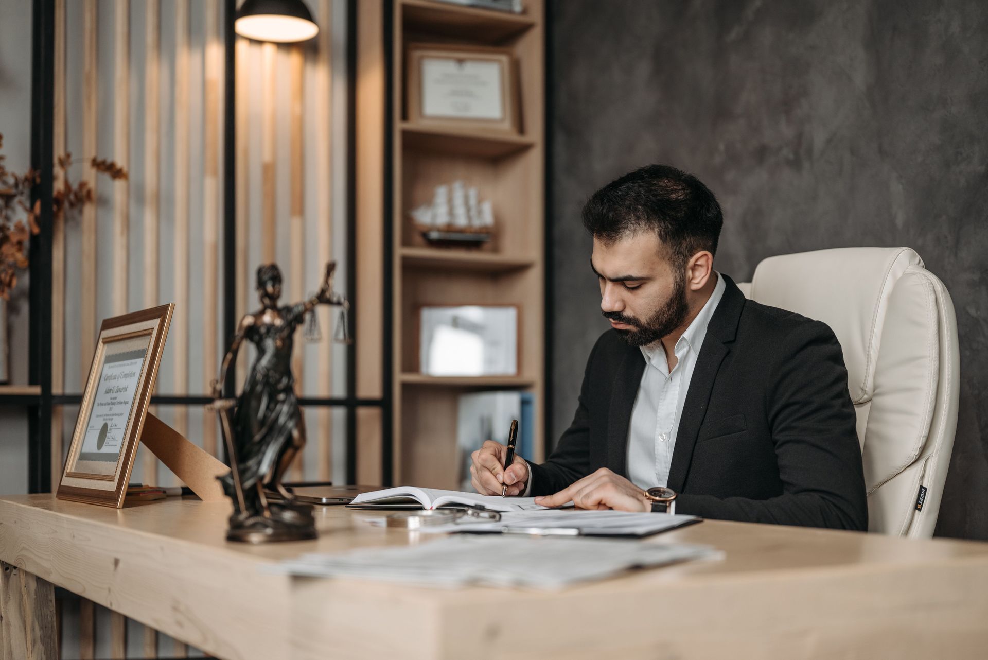 Man in a black suit writing at a desk in a modern office with shelves and a lamp.