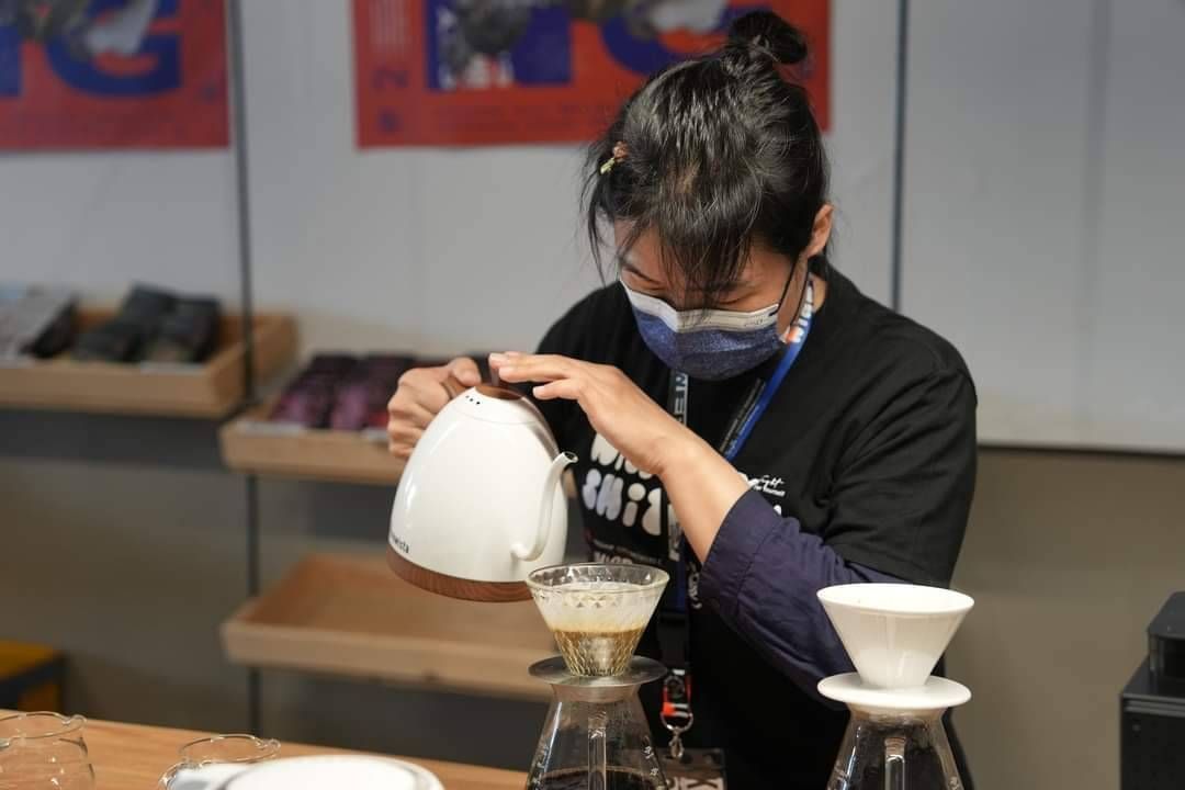 一名戴著口罩和圍裙的人正在將白色水壺中的咖啡倒入玻璃手沖咖啡器具中。