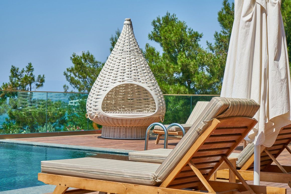 Poolside scene with wicker pod chair, lounge chairs, and closed umbrellas. Blue water and trees in the background.
