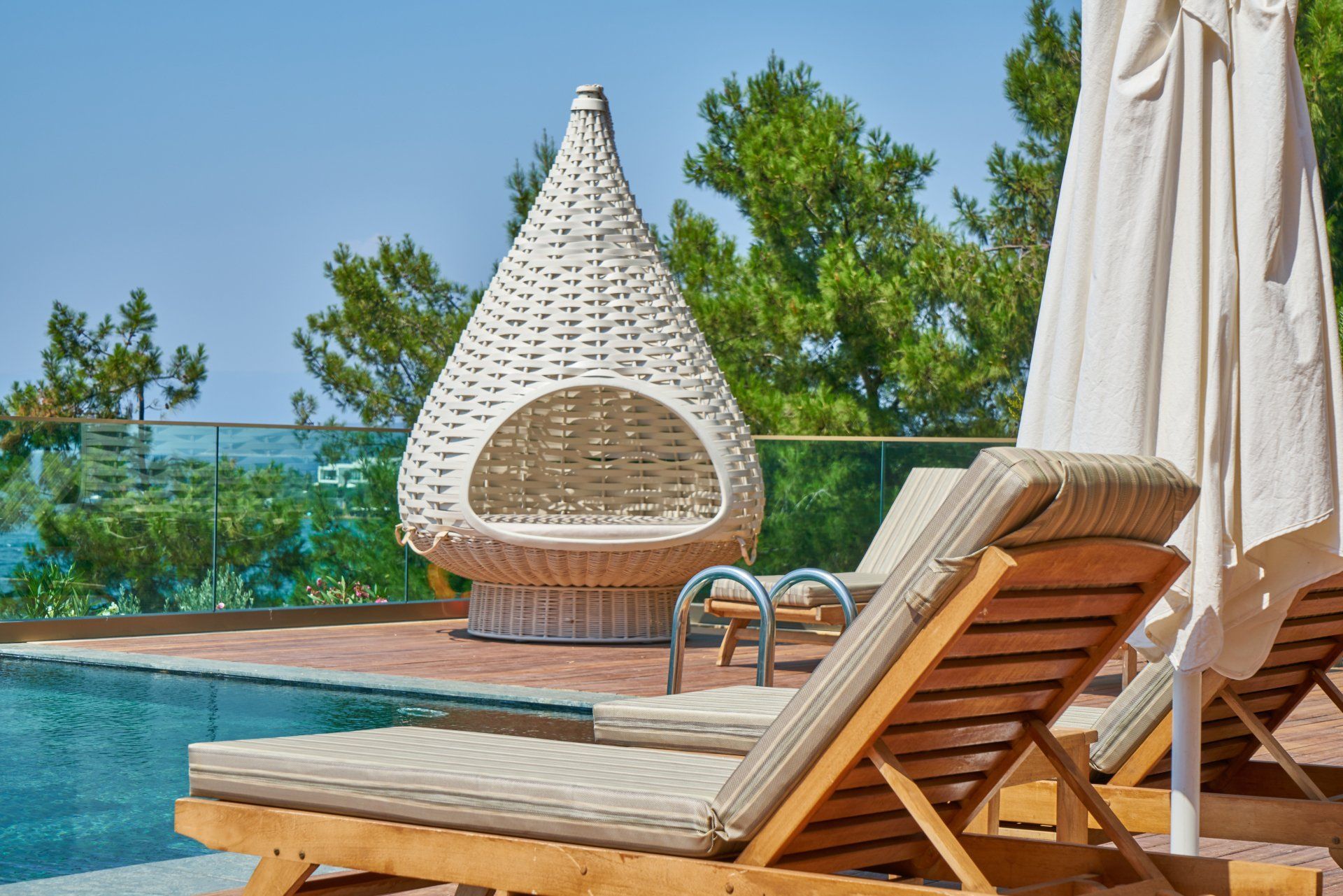 Poolside scene with wicker pod chair, lounge chairs, and closed umbrellas. Blue water and trees in the background.