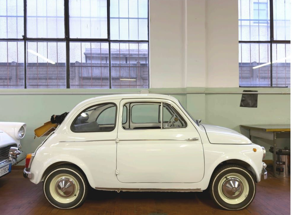 Auto compatta bianca d'epoca parcheggiata all'interno di un edificio, di fronte a grandi finestre.