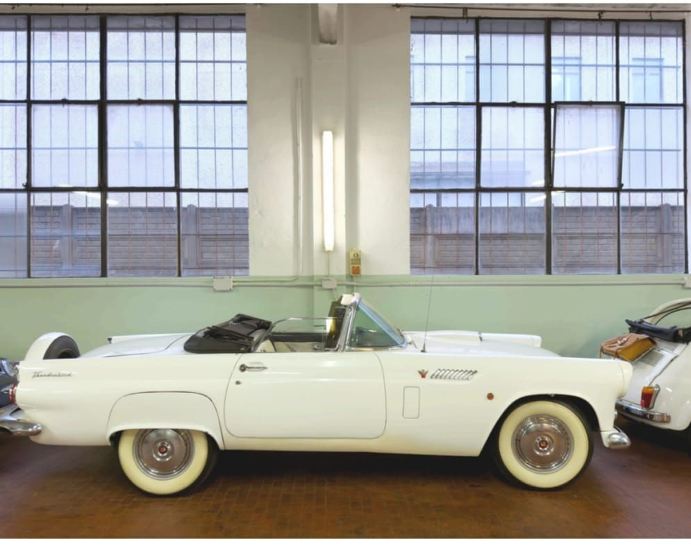 Un'auto decappottabile d'epoca bianca è esposta all'interno, accanto a un'altra auto d'epoca, davanti a grandi vetrate.