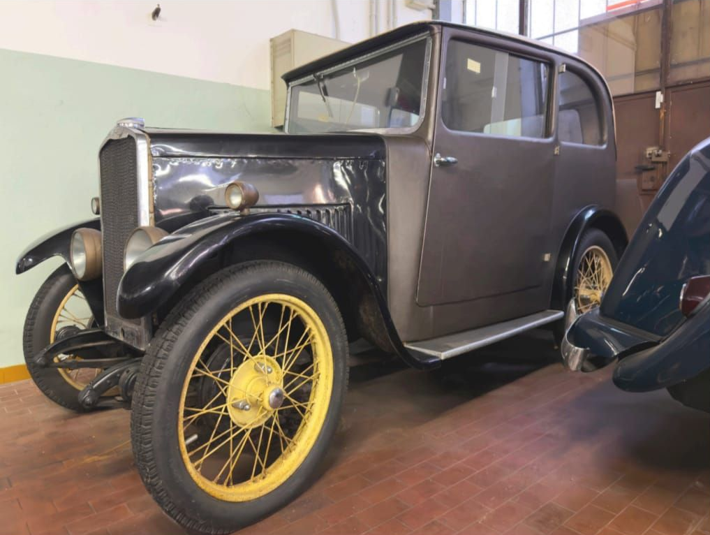 Coupé vintage grigia con cerchi a raggi gialli esposta in interni su un pavimento di legno.