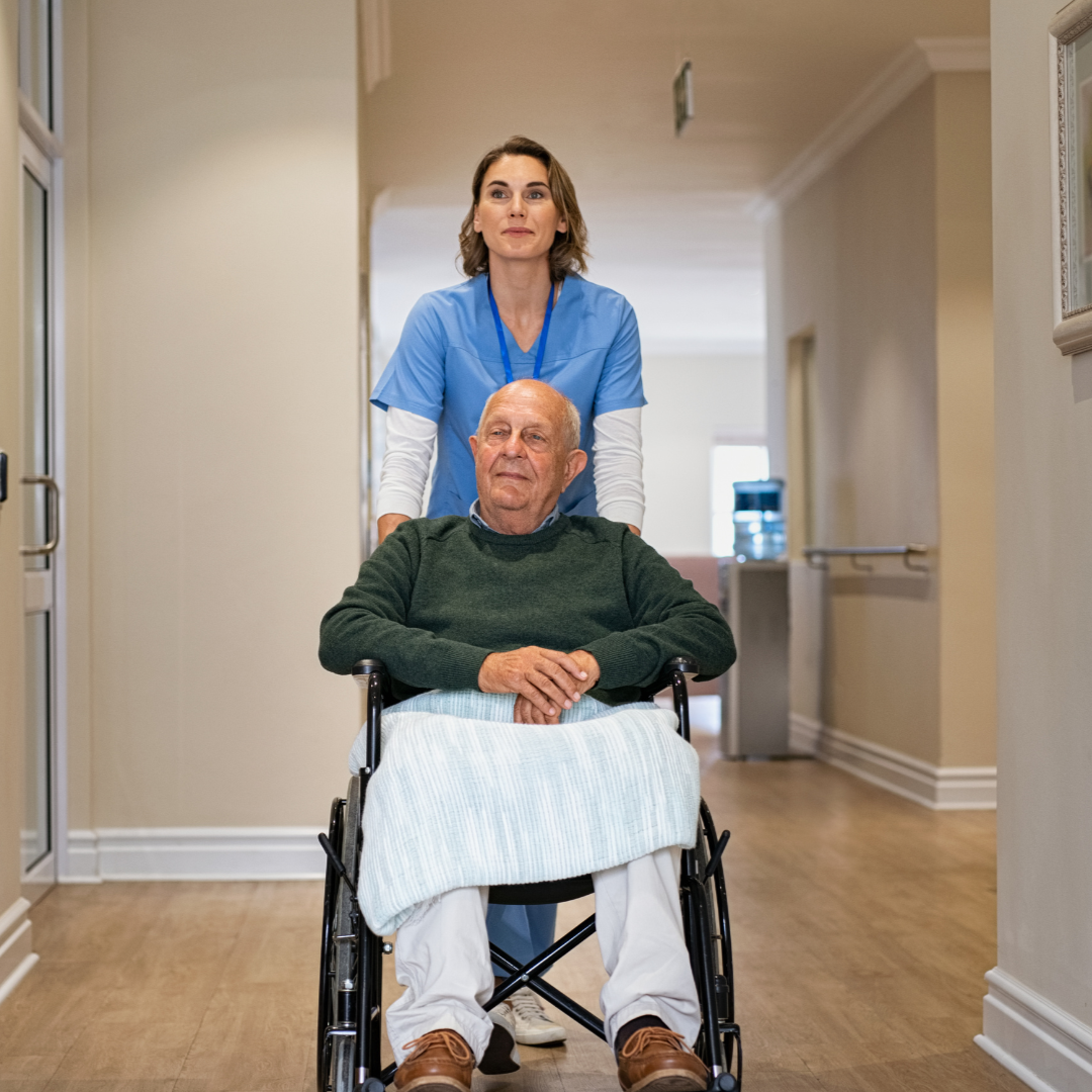 A nurse in blue scrubs pushes an elderly man in a wheelchair down a brightly lit hallway.