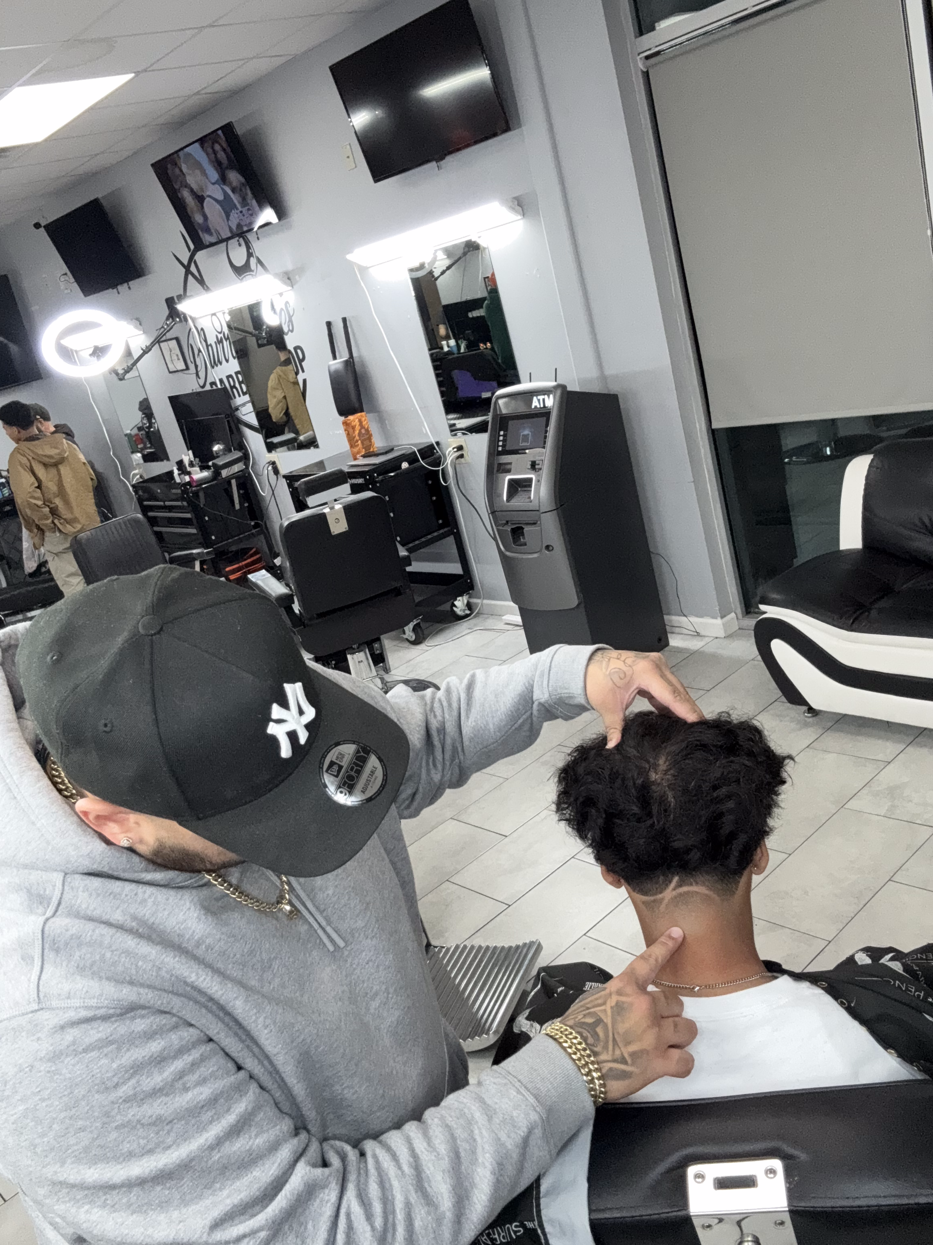 A barber in a gray hoodie and New York Yankees hat styles a client's hair in a brightly lit barbershop.
