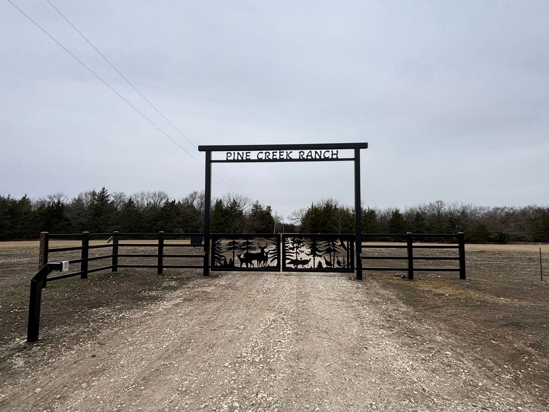 Gravel driveway leading to a gated entrance with a sign and fenced fields under an overcast sky