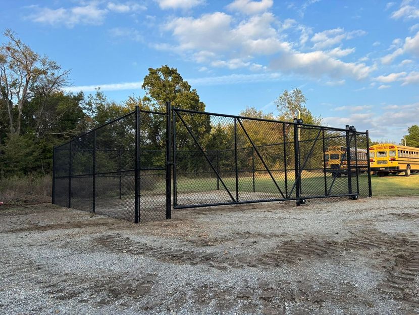 Black chain-link fenced enclosure on gravel lot with trees and yellow school buses in the background