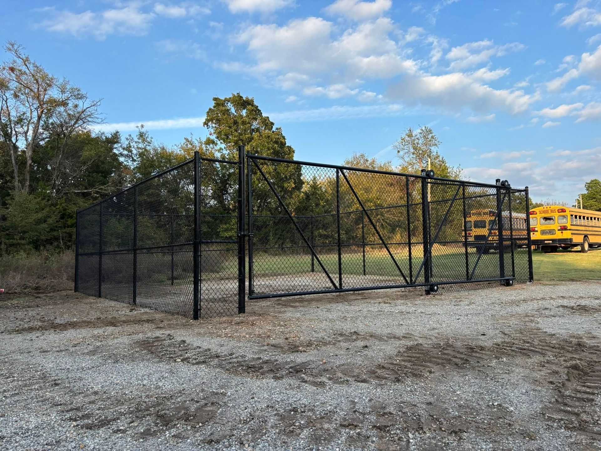 Black chain-link fenced enclosure on a gravel lot, with trees and yellow school buses in the background.