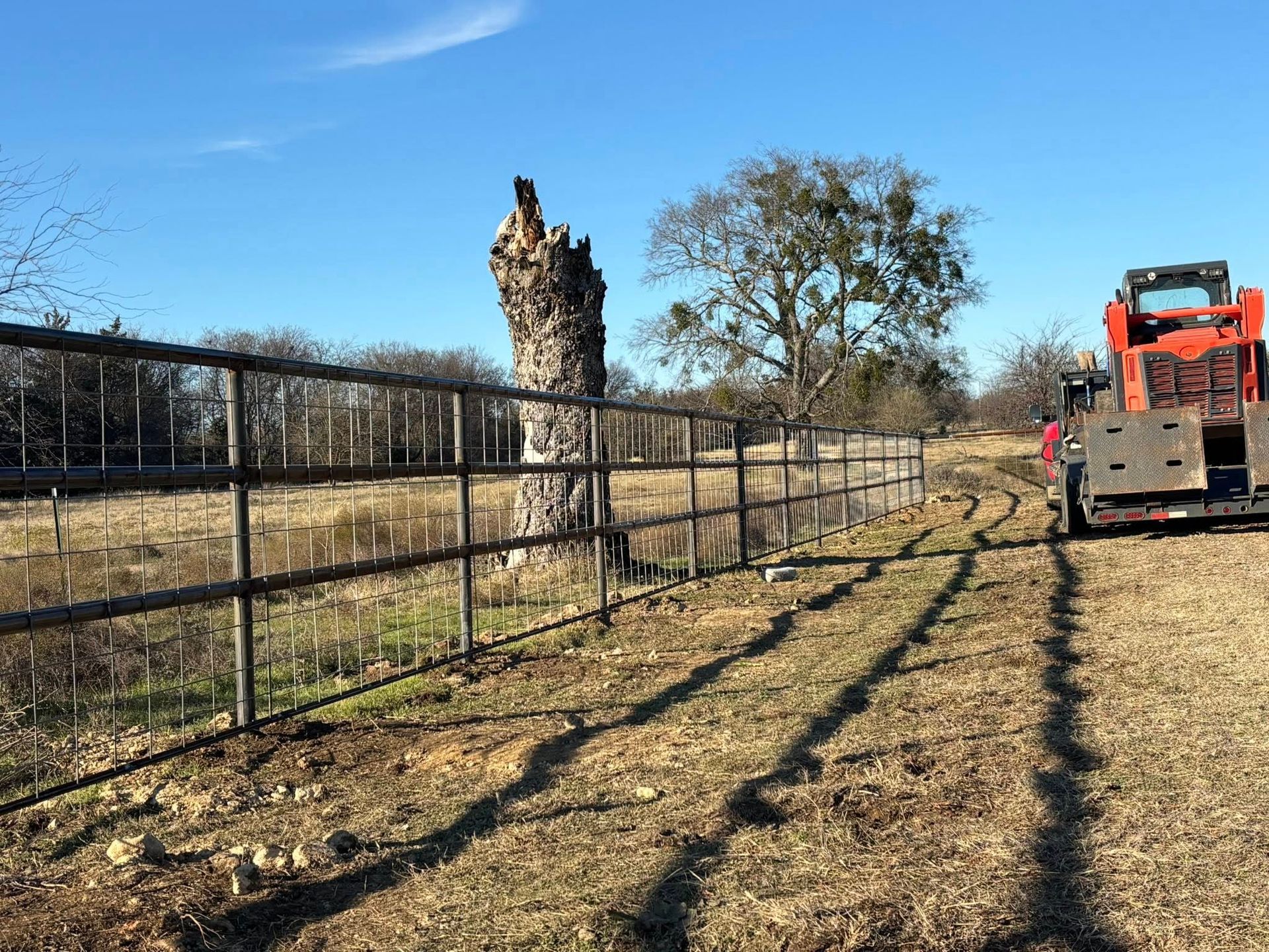 Rural pasture with a fence, leafless tree, stone stump, and an orange tractor under a clear blue sky