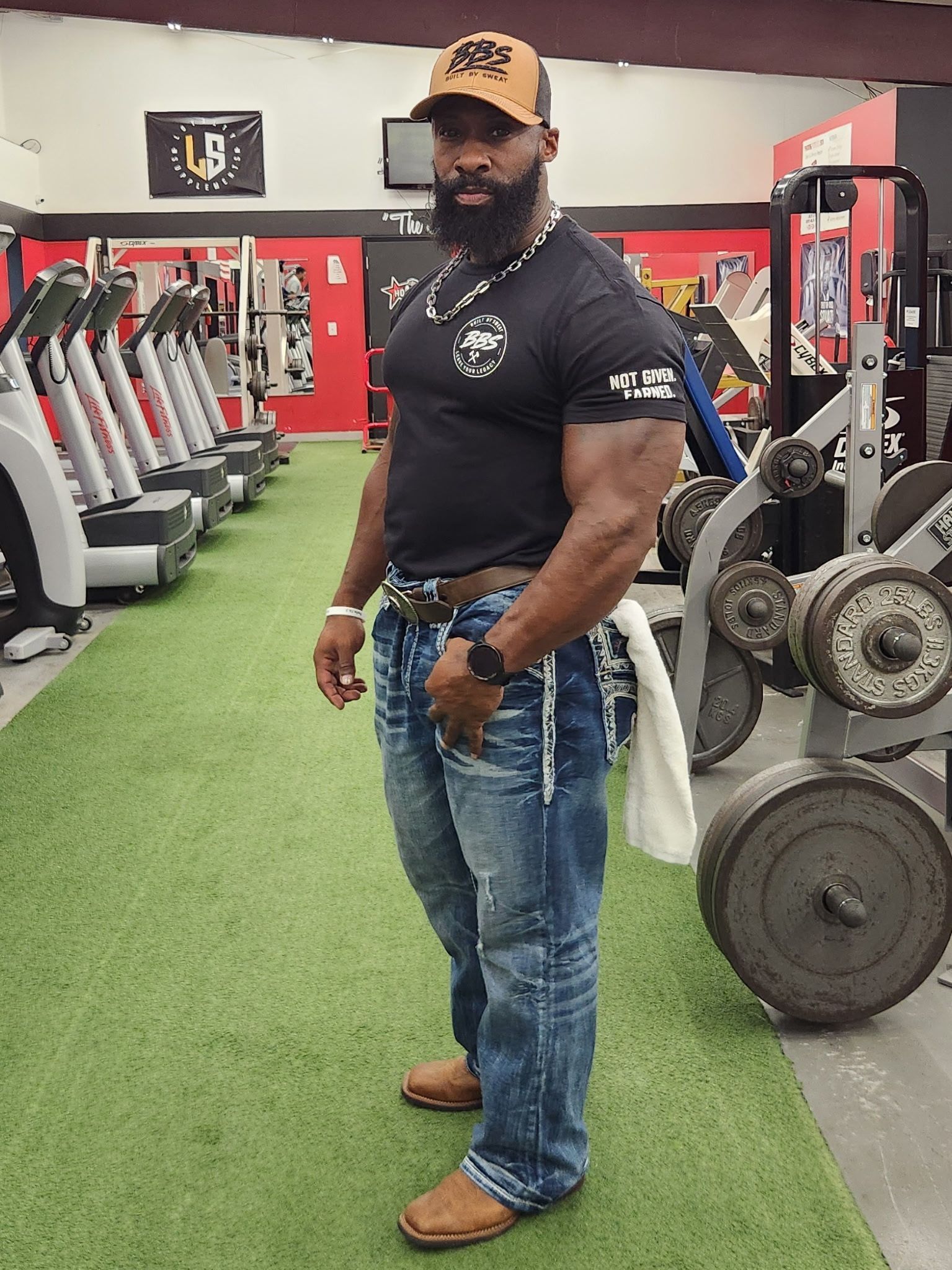 Man standing in a gym aisle beside exercise machines, wearing a black shirt, jeans, and a tan cap.