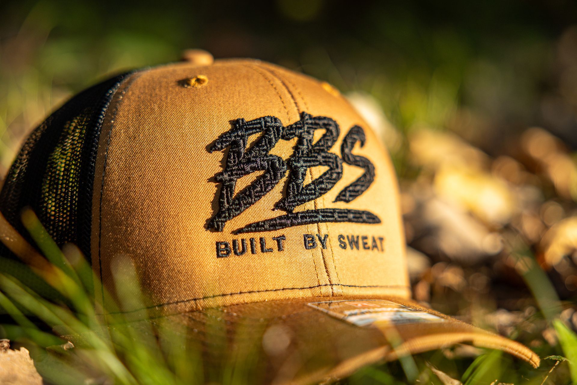 Tan mesh cap with black “BBS” logo and “BUILT BY SWEAT” text in sunlight among grass