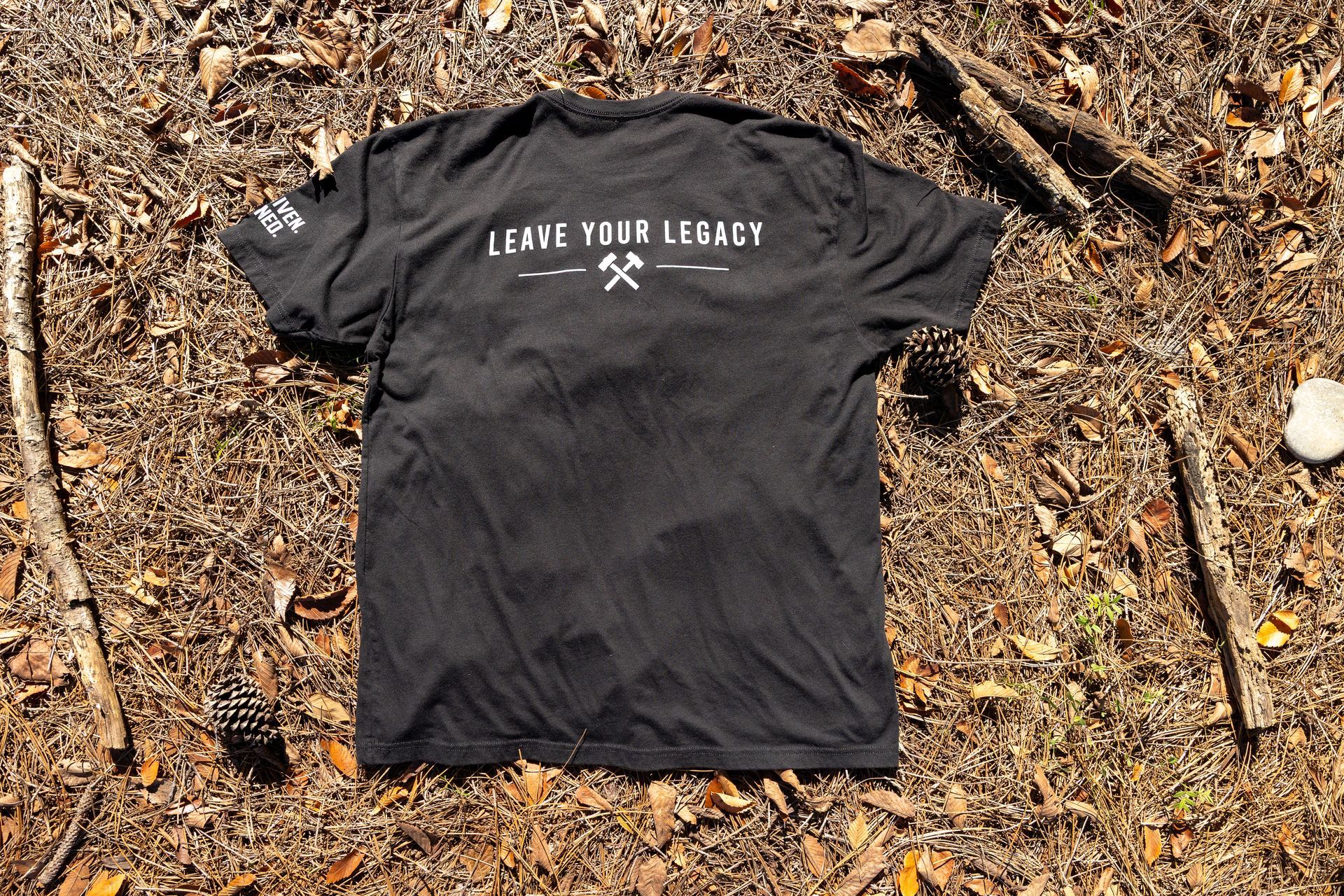 Black T-shirt with white circular BBS logo laid on pine needles outdoors
