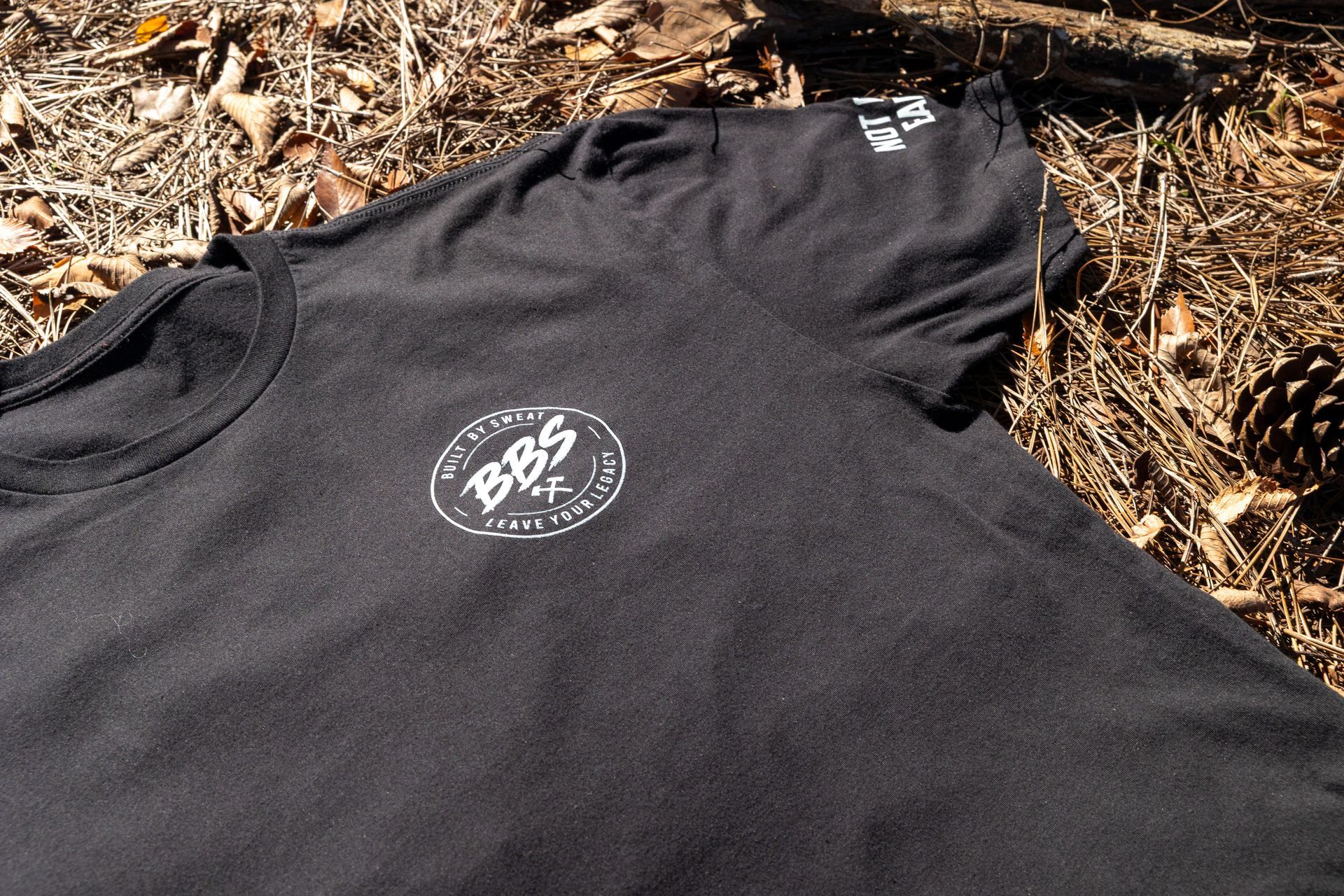 Black T-shirt with white circular BBS logo laid on pine needles outdoors