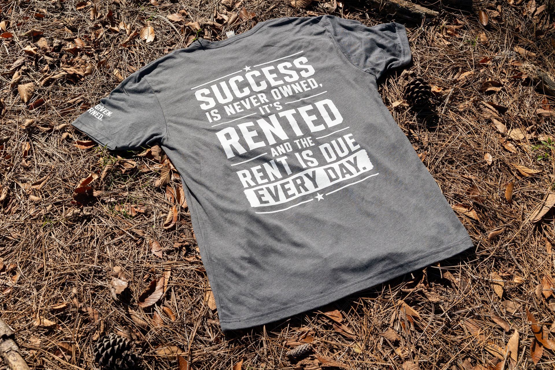 Gray T-shirt with white text lying on brown leaves outdoors