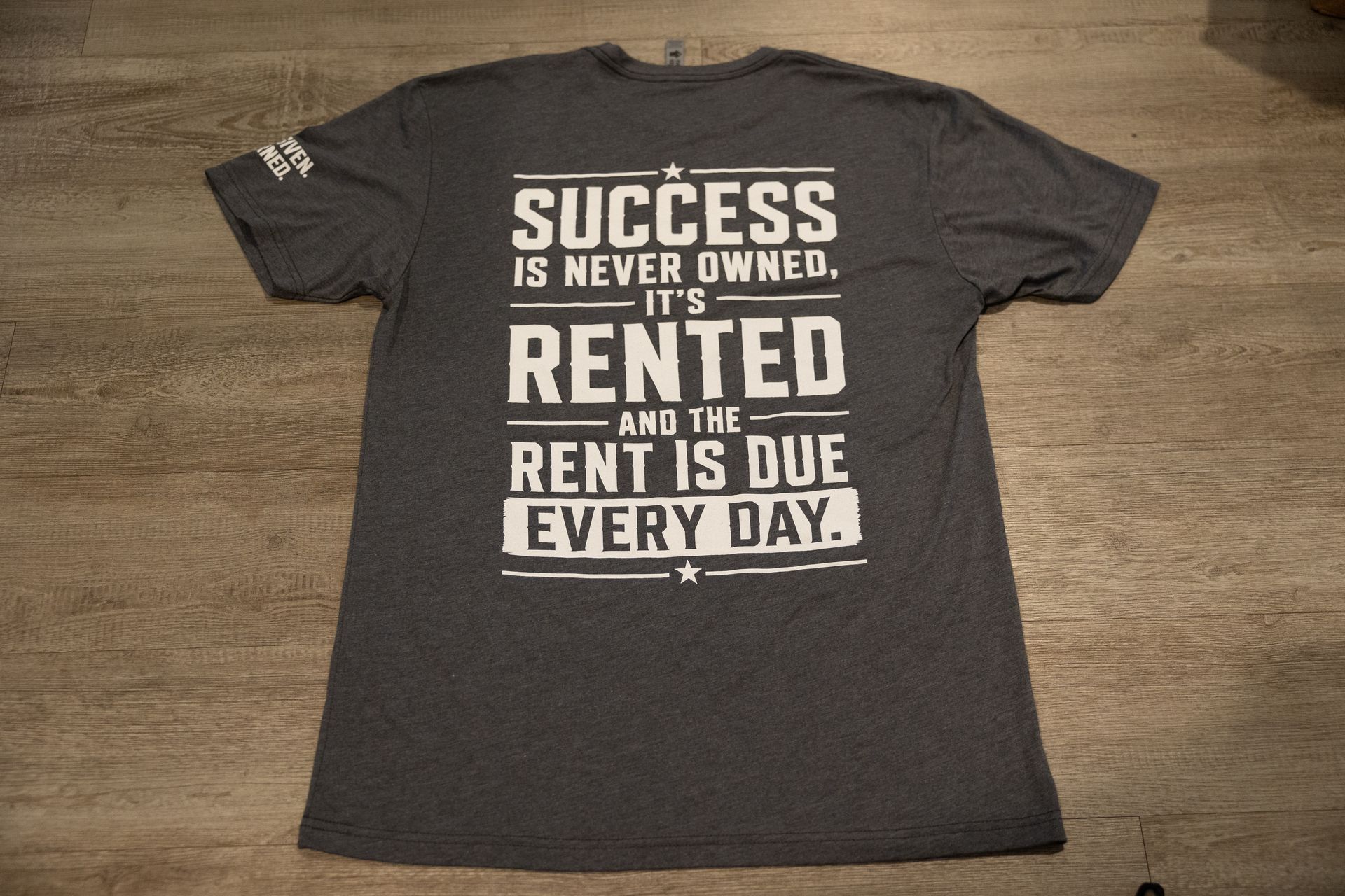 Gray T-shirt on dirt with bold black text: “SUCCESS IS NEVER OWNED, IT'S RENTED AND THE RENT IS DUE EVERY DAY.”
