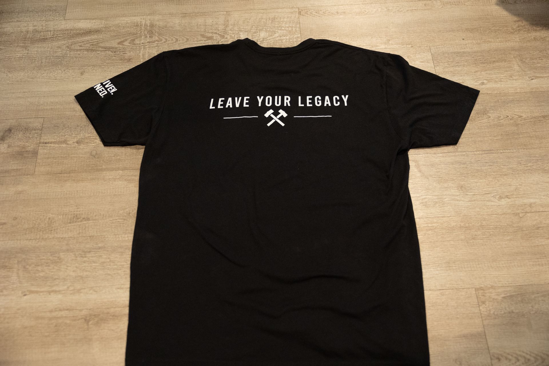 Black T-shirt with “LEAVE YOUR LEGACY” text and crossed tools graphic laid flat on a wooden floor