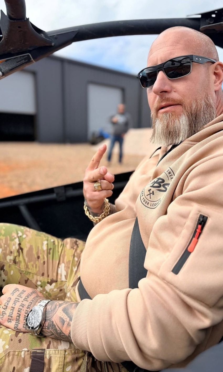 Man in sunglasses and tan hoodie seated in an open vehicle, giving a peace sign outdoors.