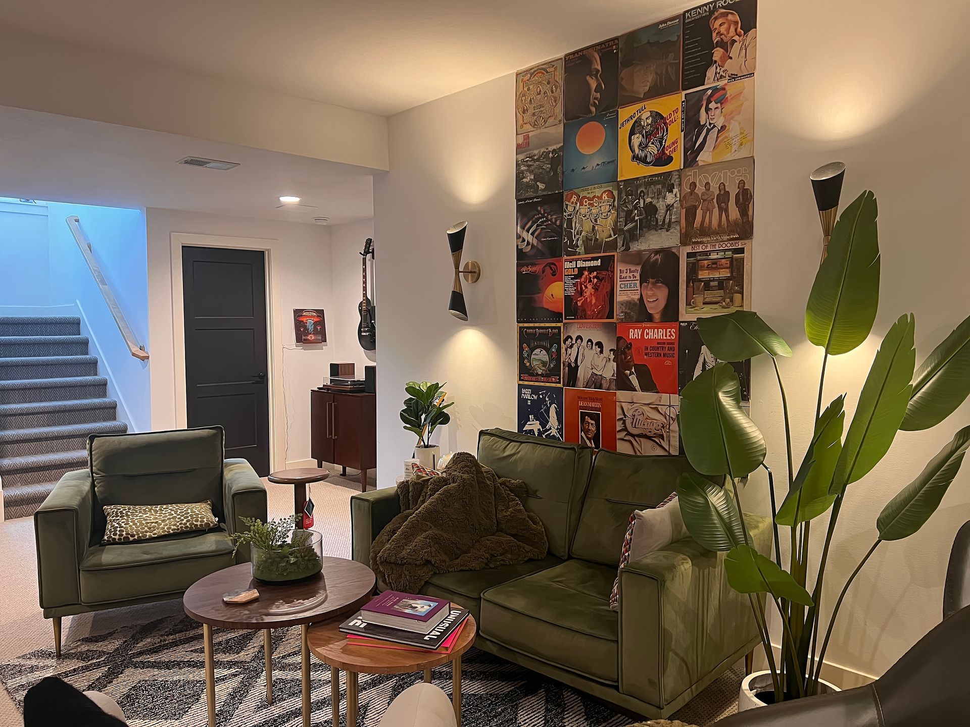 Cozy living room with green sofa, armchair, potted plants, wall art, and stairway in the background