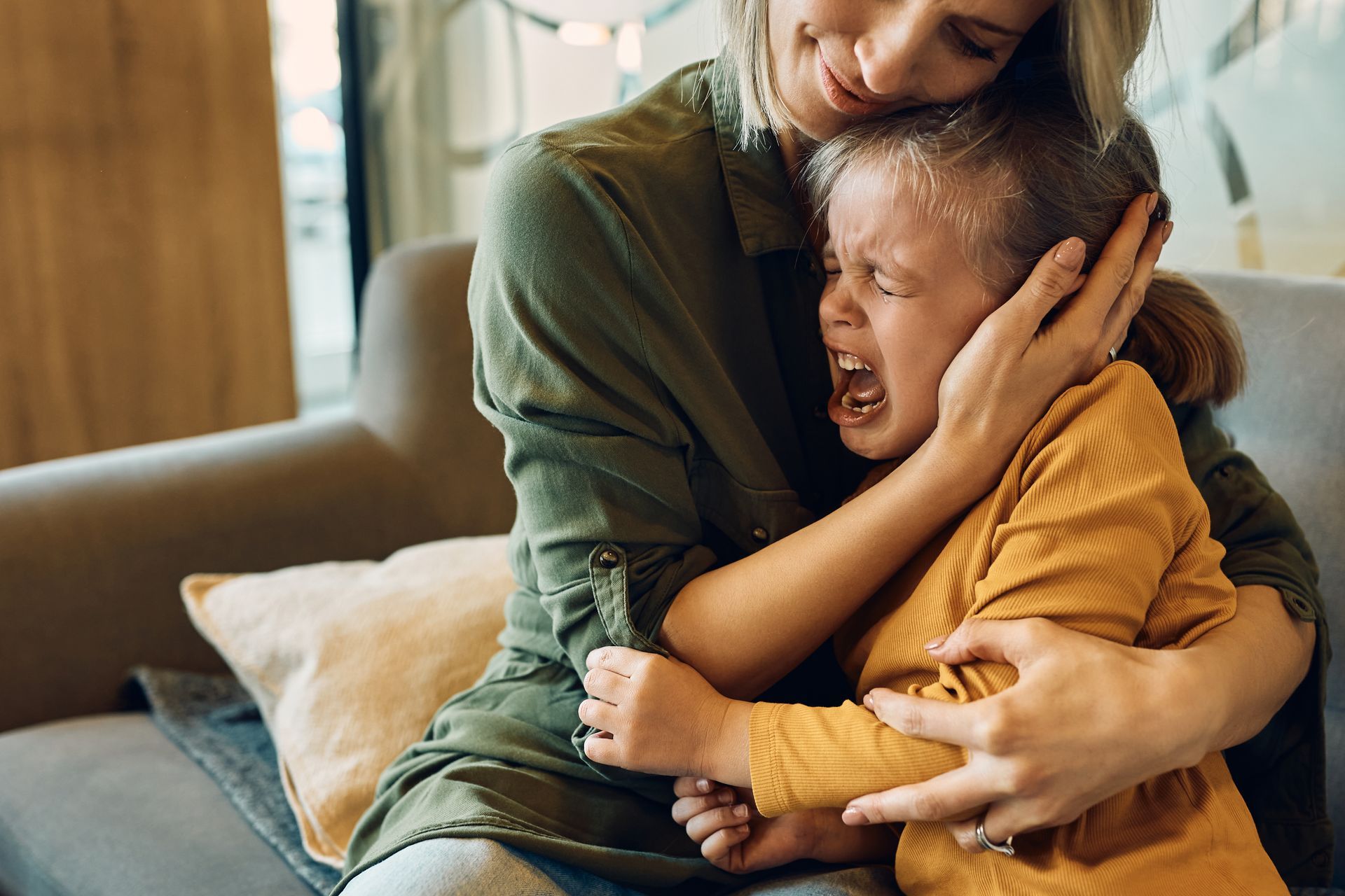 Mother comforting crying child; arms wrapped around, indoor setting.