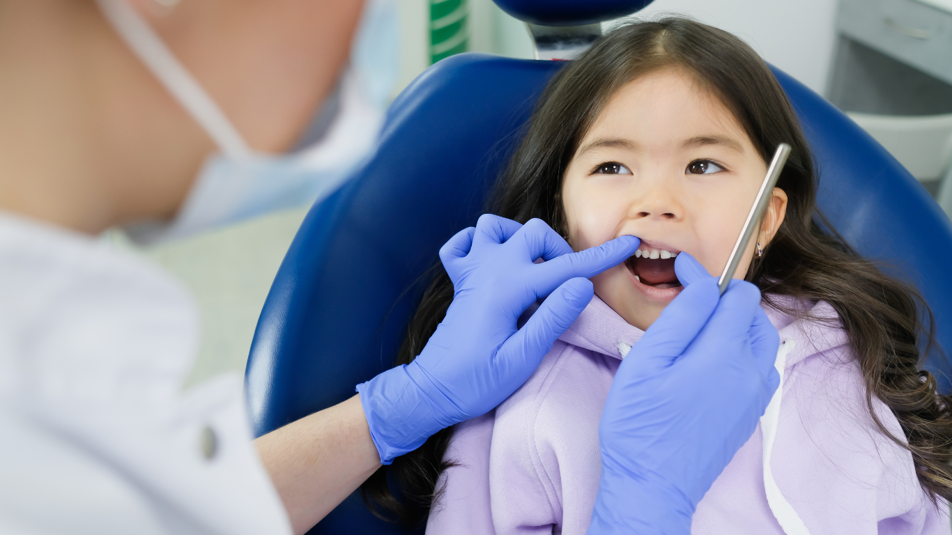 A dentist in blue gloves examines a child's teeth in a clinical setting.