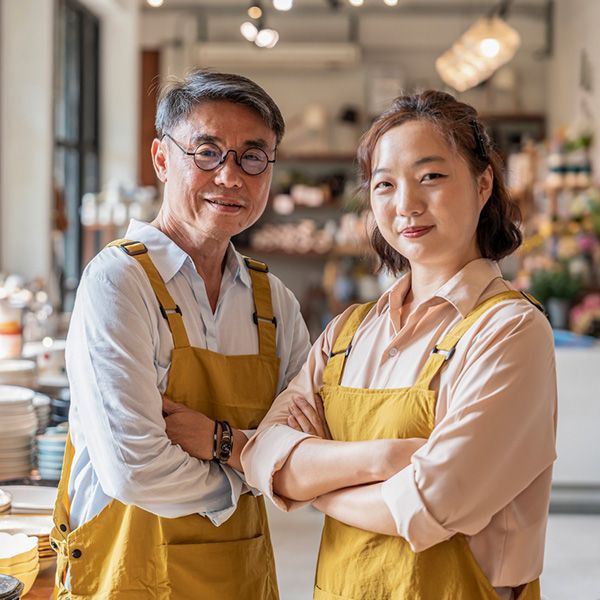 Two shop keepers in front of homewares, a man and a woman, both wearing yellow aprons.