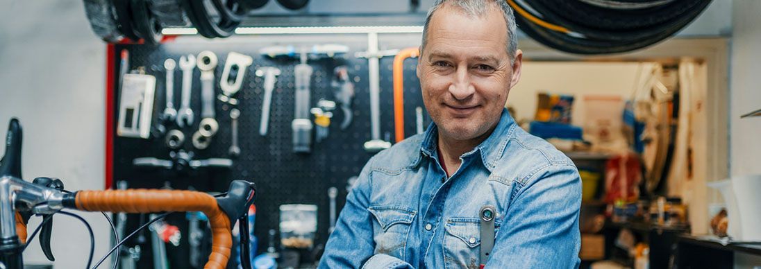 The male owner of a a bike shop looking at the camera. He has grey hair and a blue shirt and is surrounded by tools.