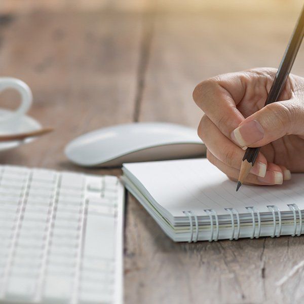 Keyboard and mouse on a desk. Hand with pen hovering over a notebook.