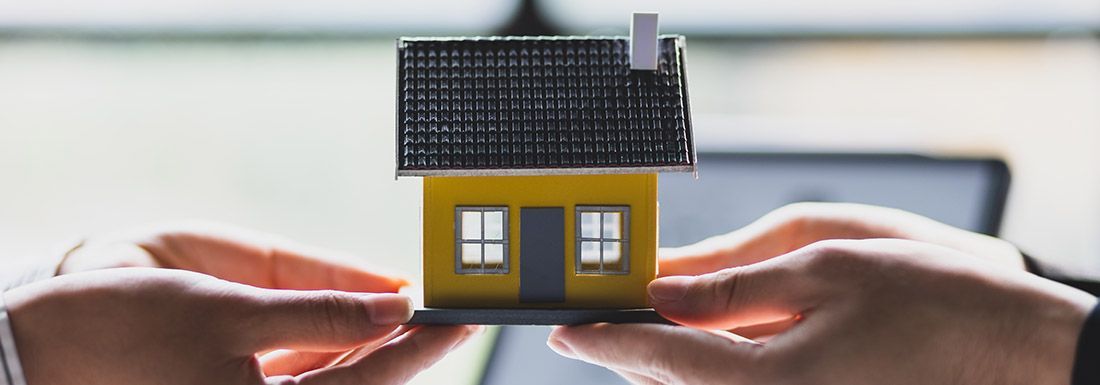 An image of two hands exchanging a model of a home. The home is yellow.
