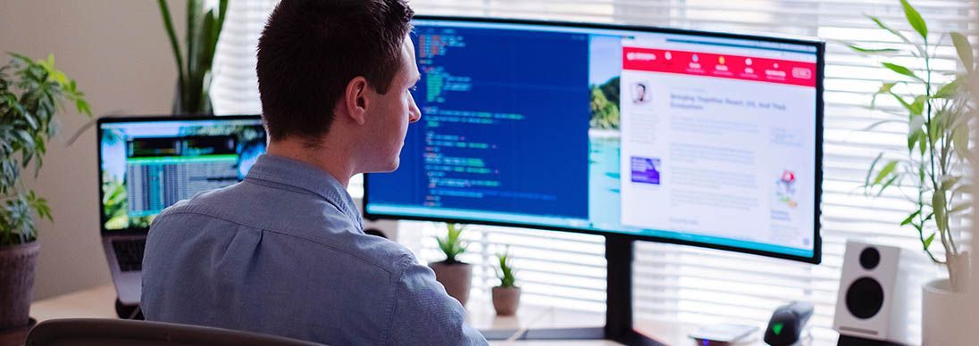 A man working from home facing two screens. he is wearing a blue shirt and has short dark hair.