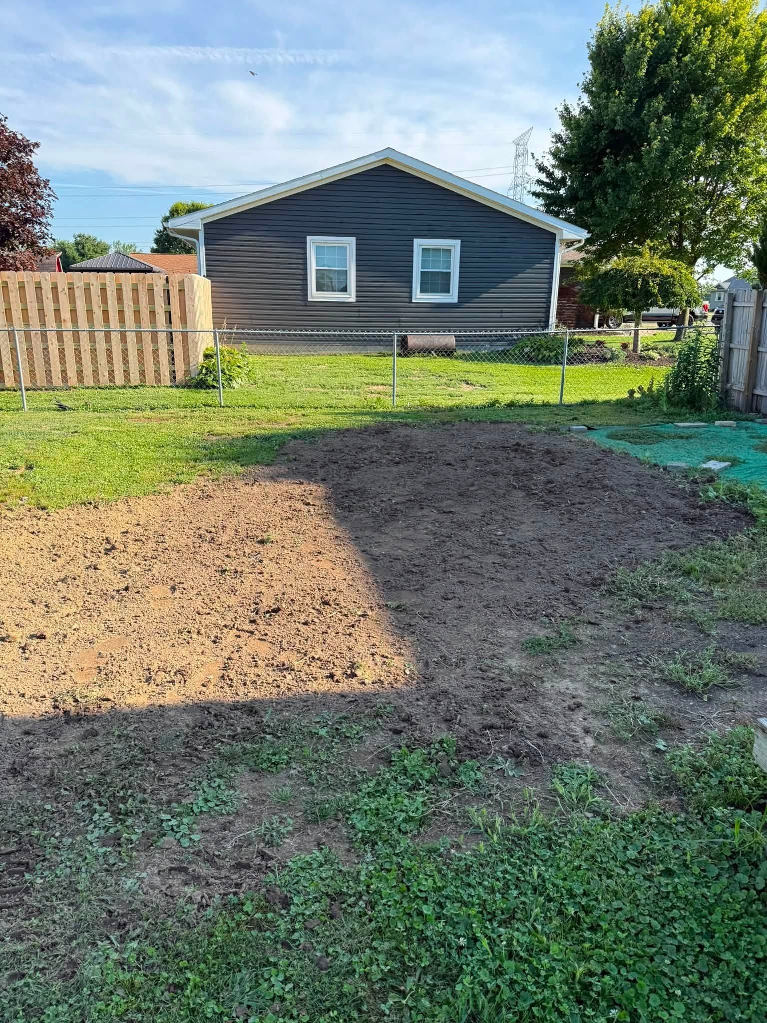 Backyard with a dirt patch, green grass, wooden fence, and gray shed under a blue sky.