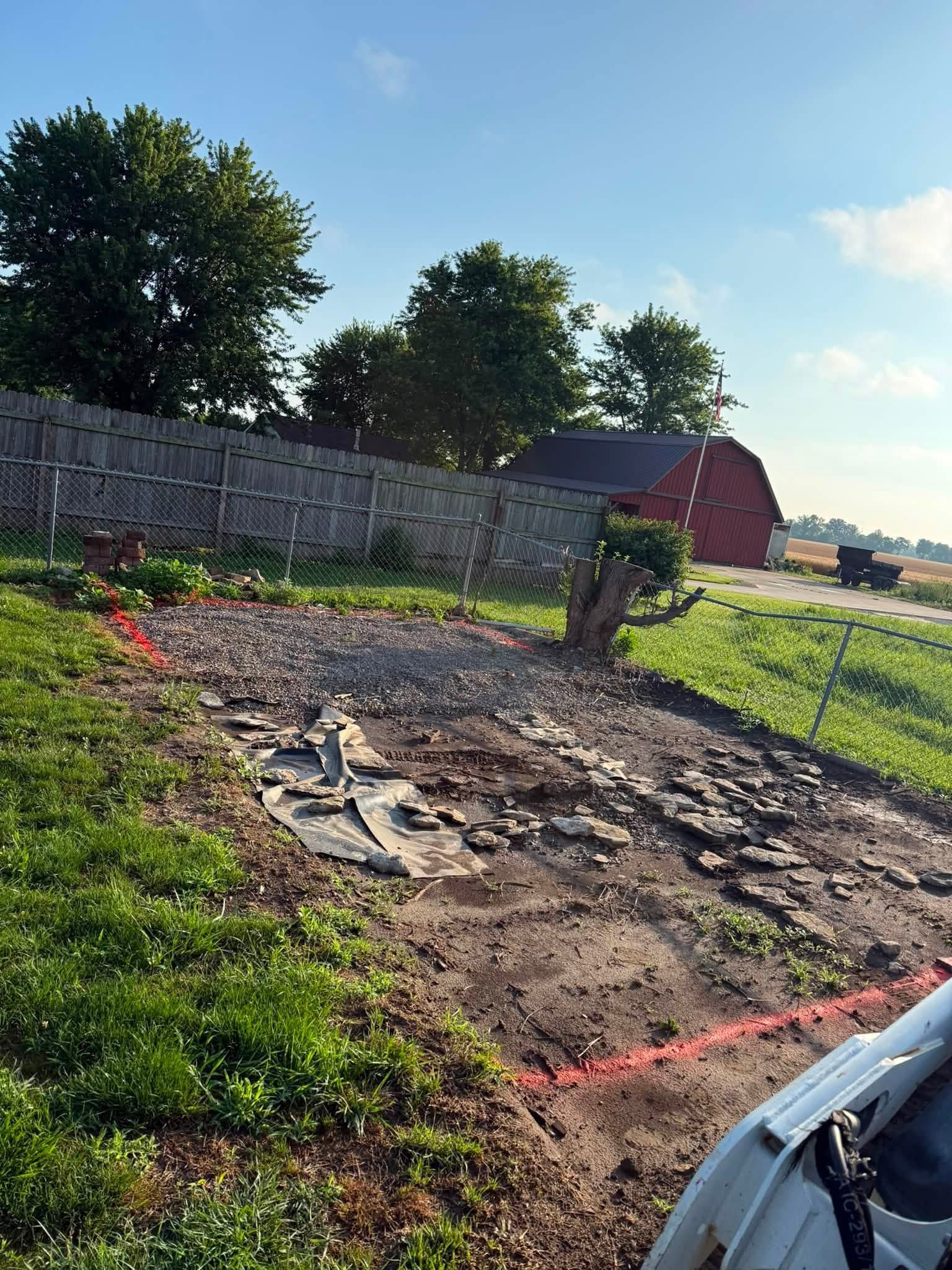 Sunny backyard garden bed with tilled soil, grass edges, fence, trees, and a red shed in the distance