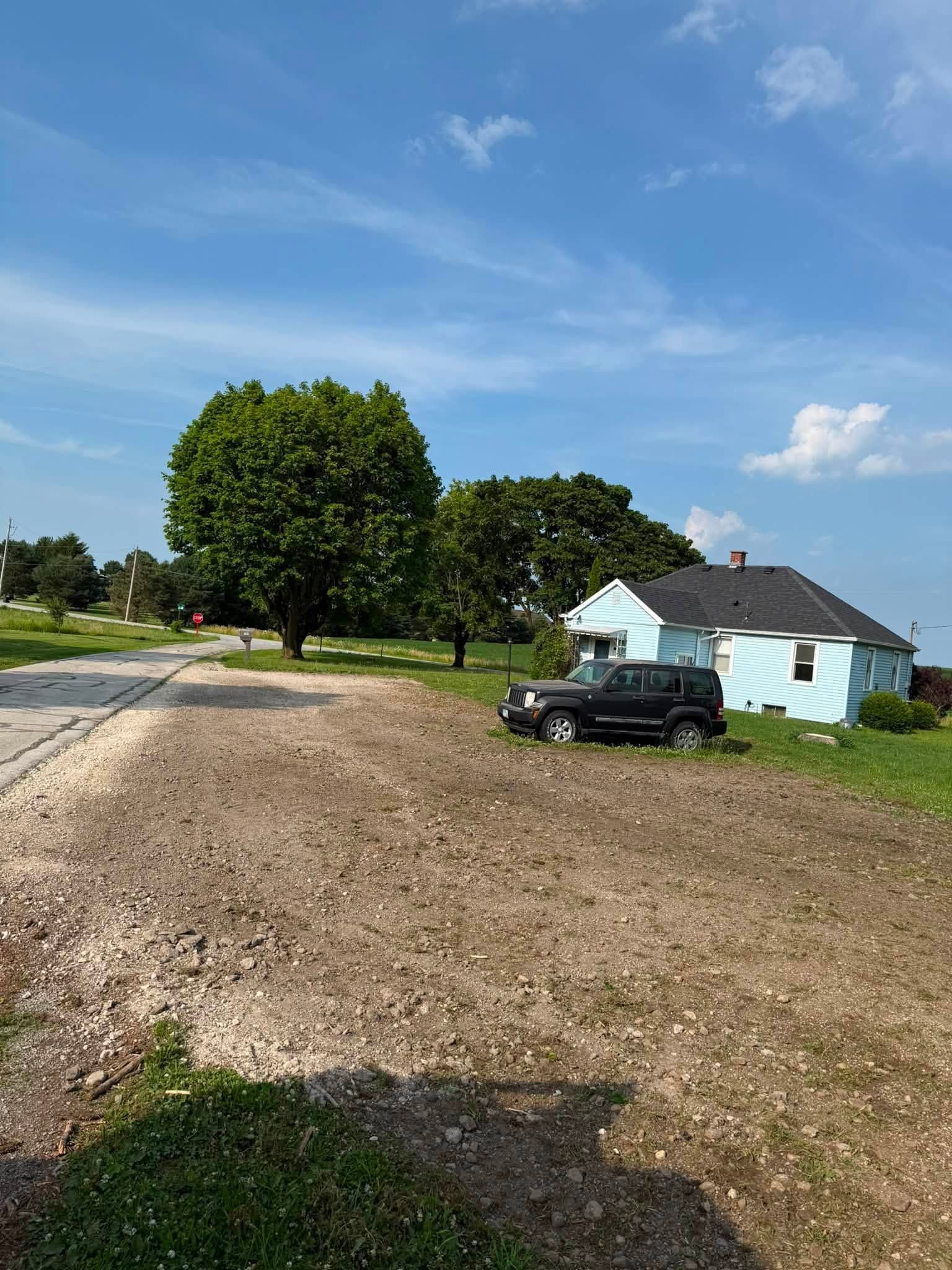 Gravel driveway beside a light blue house, with a black SUV and large trees under a sunny sky
