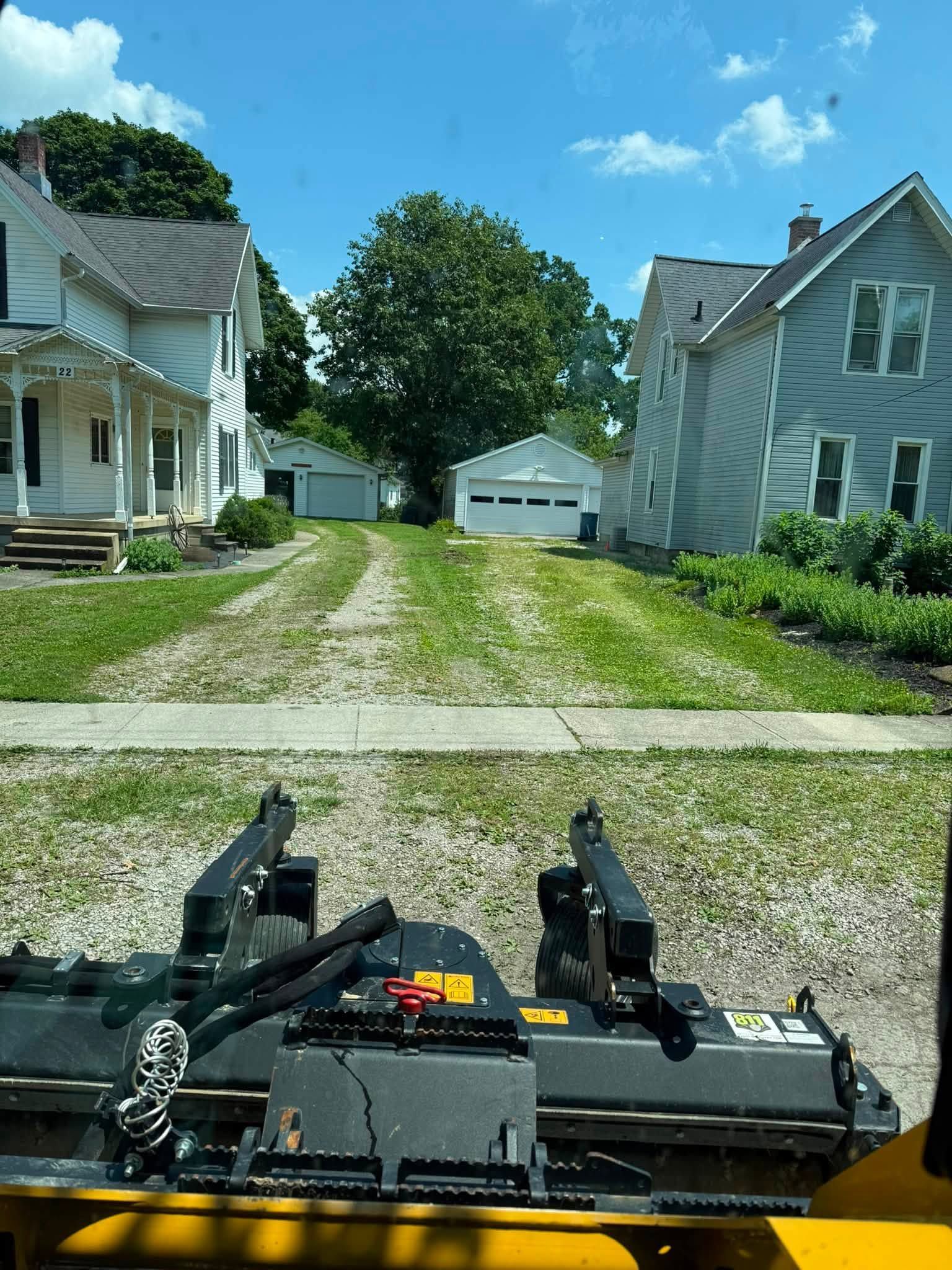 First-person view from a lawn mower on a gravel driveway between white houses, heading toward a garage