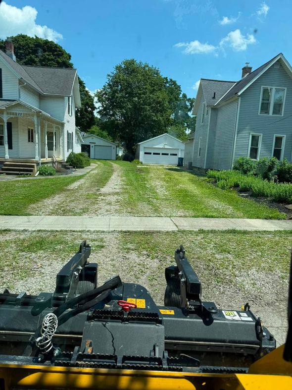 Riding mower view down a gravel driveway between white houses toward a garage on a sunny day