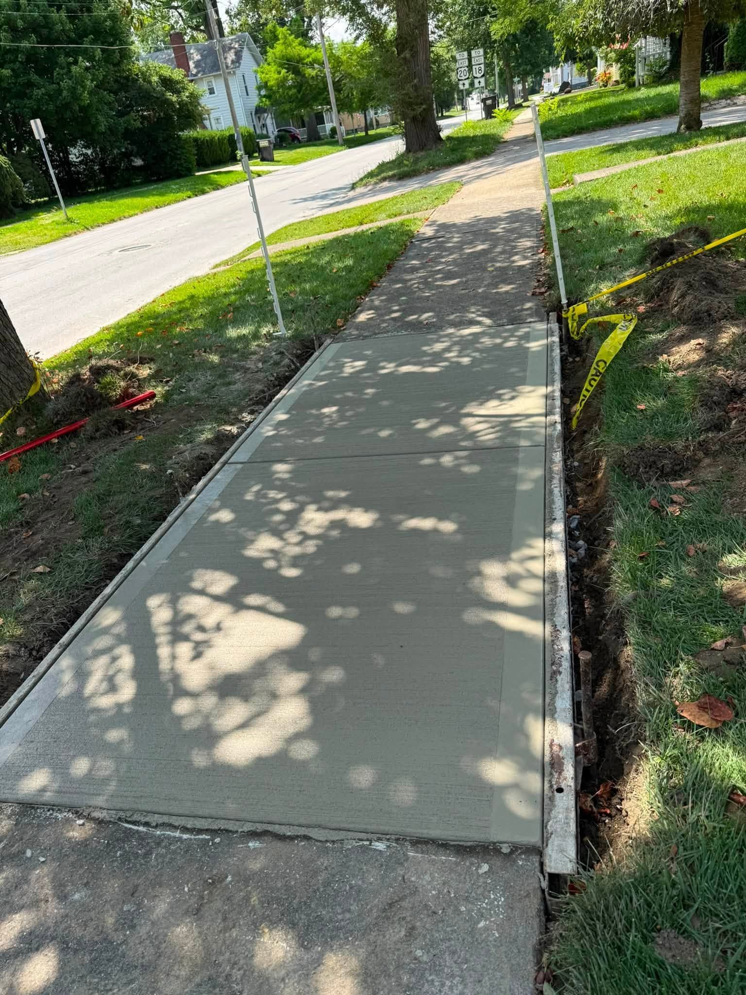 Concrete sidewalk beside a grassy roadside, with a curb and tree shadows in bright daylight