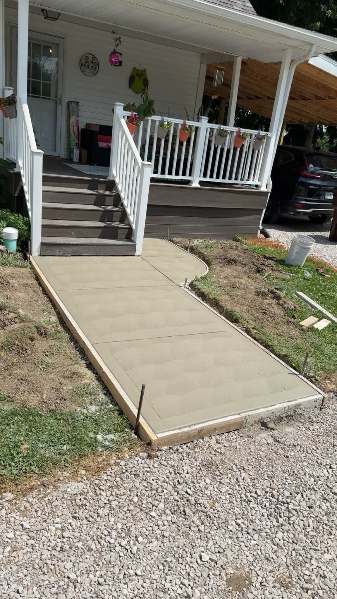 Concrete ramp leading to a porch with white railings and steps, beside a gravel driveway.