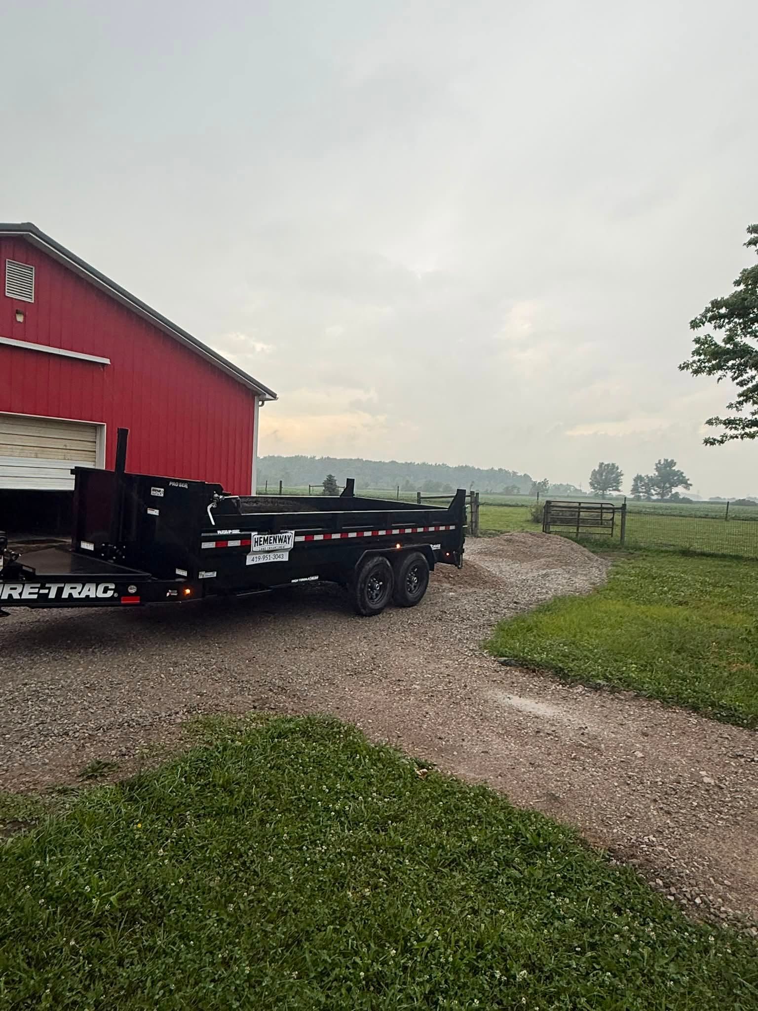 Red barn on a gravel path beside a flatbed trailer in a misty rural field.