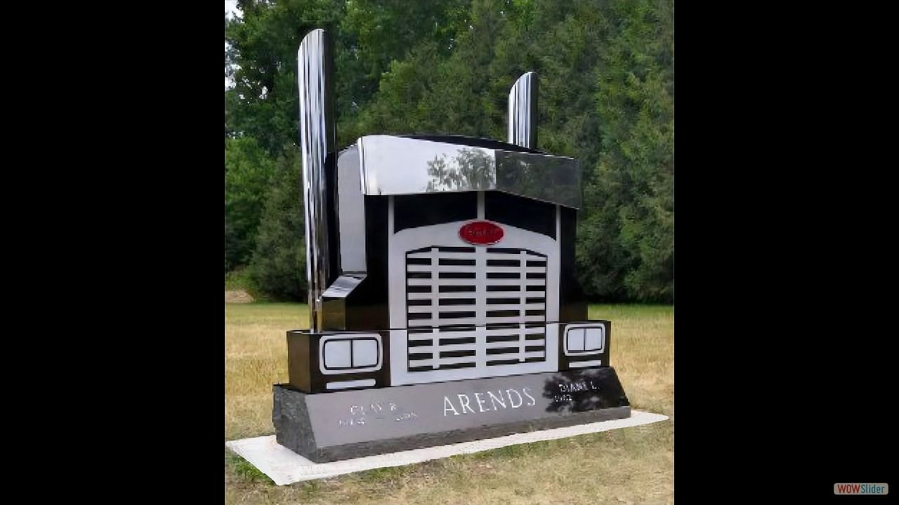 A black granite gravestone shaped like the front of a Peterbilt semi-truck with chrome-colored exhaust pipes and grille.