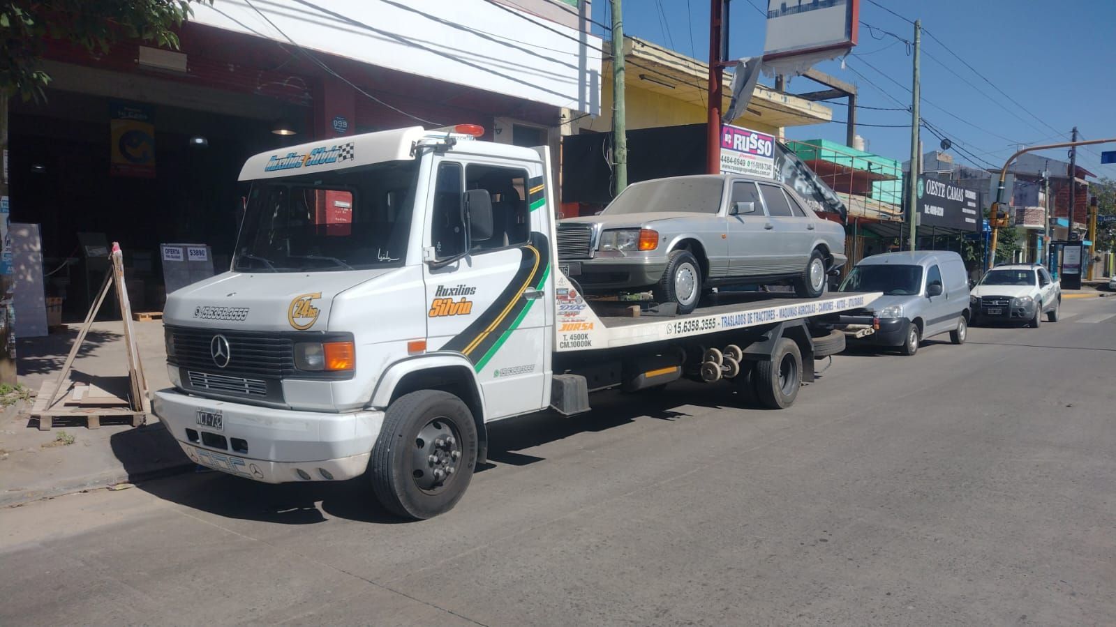 Sedán plateado sobre una grúa de plataforma azul circulando por una autopista.