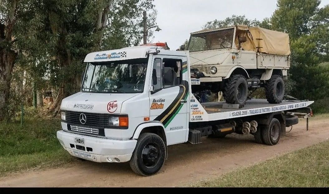 Una furgoneta Mercedes argentina azul y blanca circula por una carretera arbolada, remolcando un remolque de plataforma.