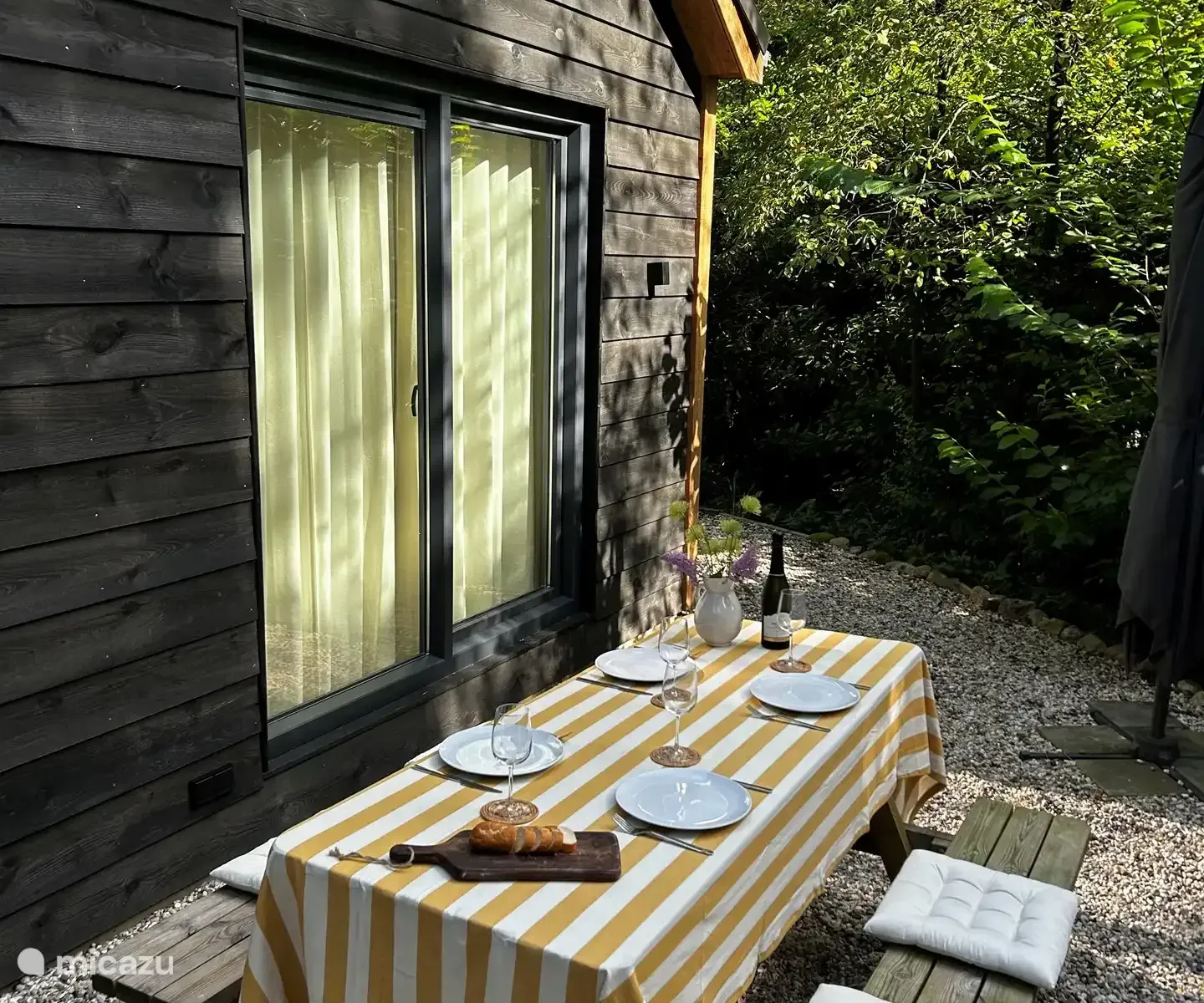 Tafel gedekt voor een maaltijd buiten een blokhut, met een geel gestreept tafelkleed, borden en wijn.