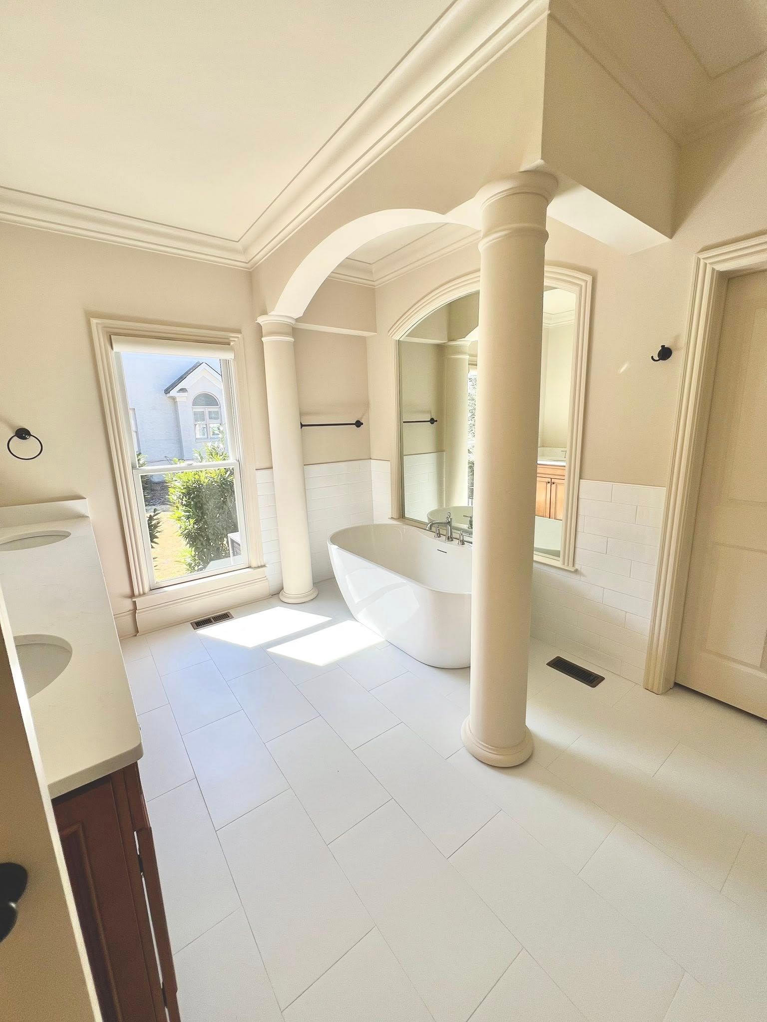 Bright cream bathroom with freestanding tub, column, tiled floor, and sunlight from the window.