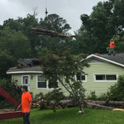 Crane lifting debris from a damaged roof while workers in orange vests stand near a lime-green house
