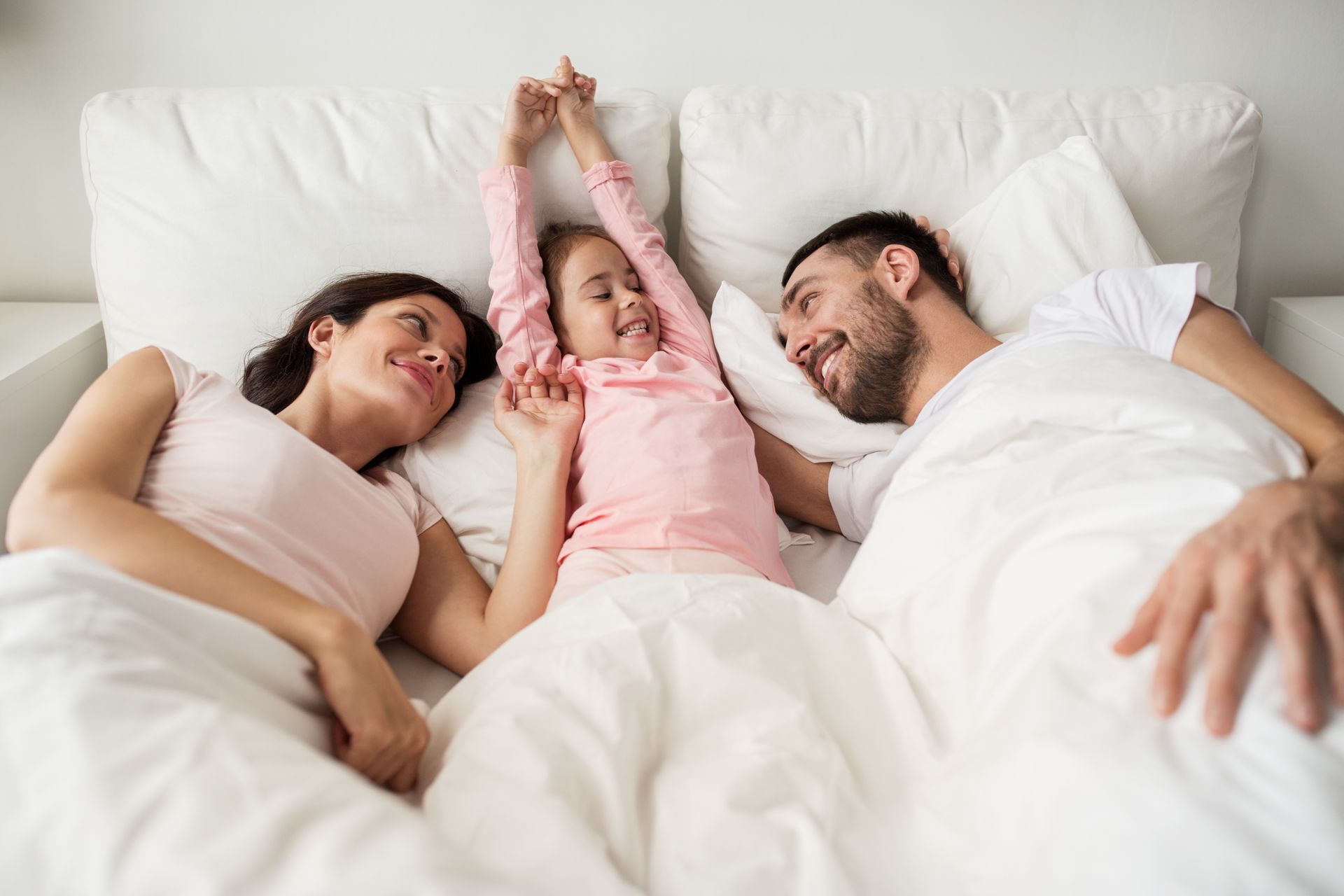 Family lying in bed with a smiling child between two adults, stretching their arms up. Family lying in bed with a smiling child between two adults, stretching their arms up.