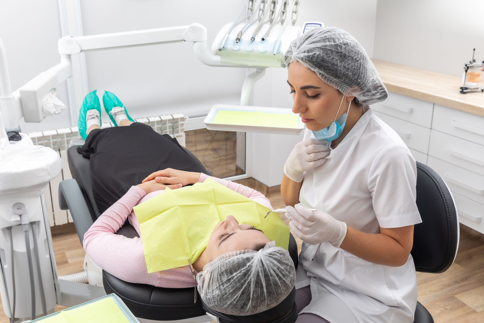 Dentist examining a patient in a dental chair with a green bib and mask in a bright clinic.