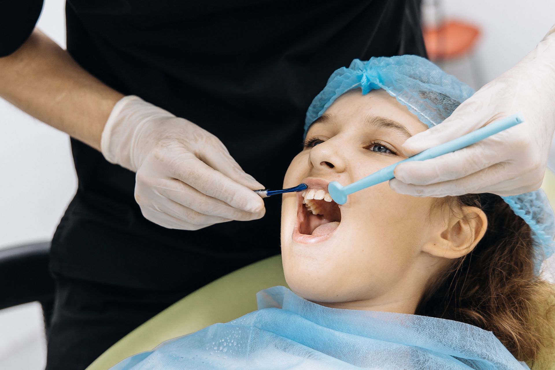 Dentist examining a child’s open mouth with dental tools, wearing gloves and a blue hair cover