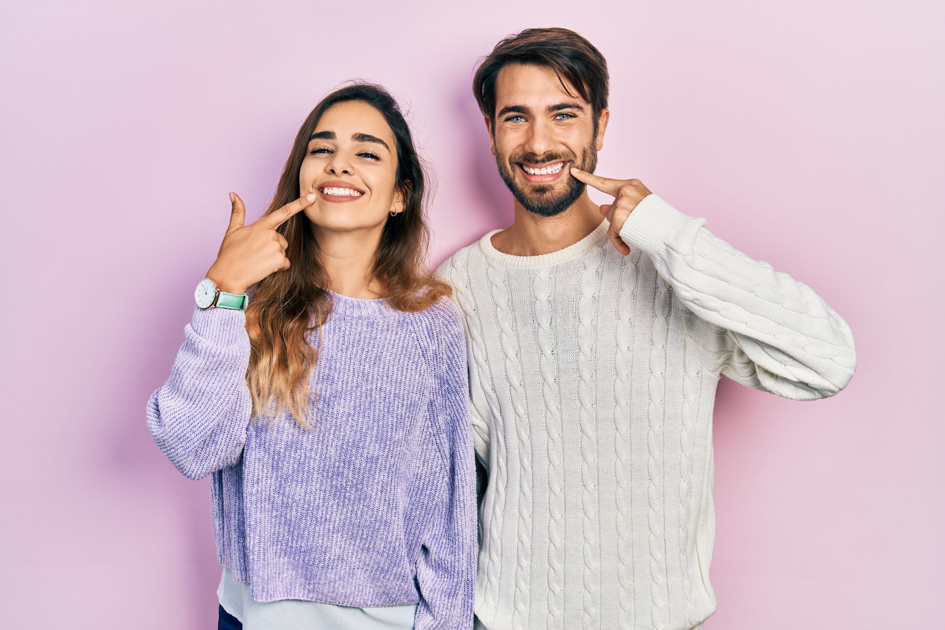 Two smiling people in cozy sweaters posing against a pink background, pointing at themselves
