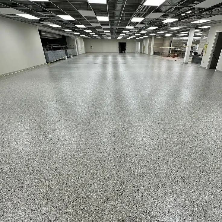 Empty fluorescent-lit warehouse with gray speckled floor and metal shelves along the walls