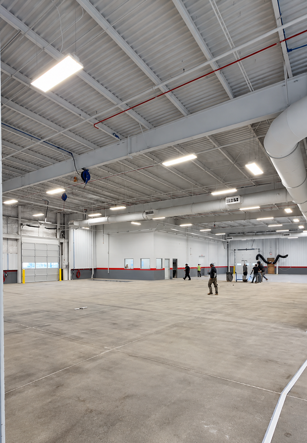 Large empty warehouse interior with a high ceiling, concrete floor, and a few people standing near the far wall