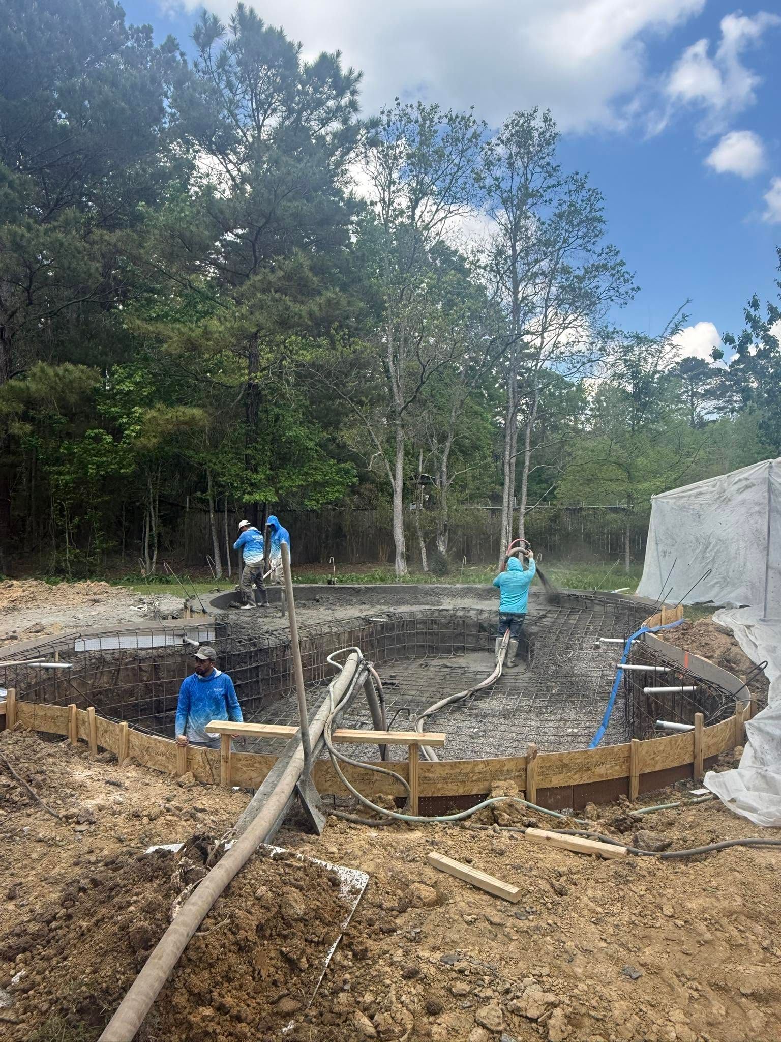 Workers building a round concrete foundation at a wooded site, with forms, rebar, and excavation visible.
